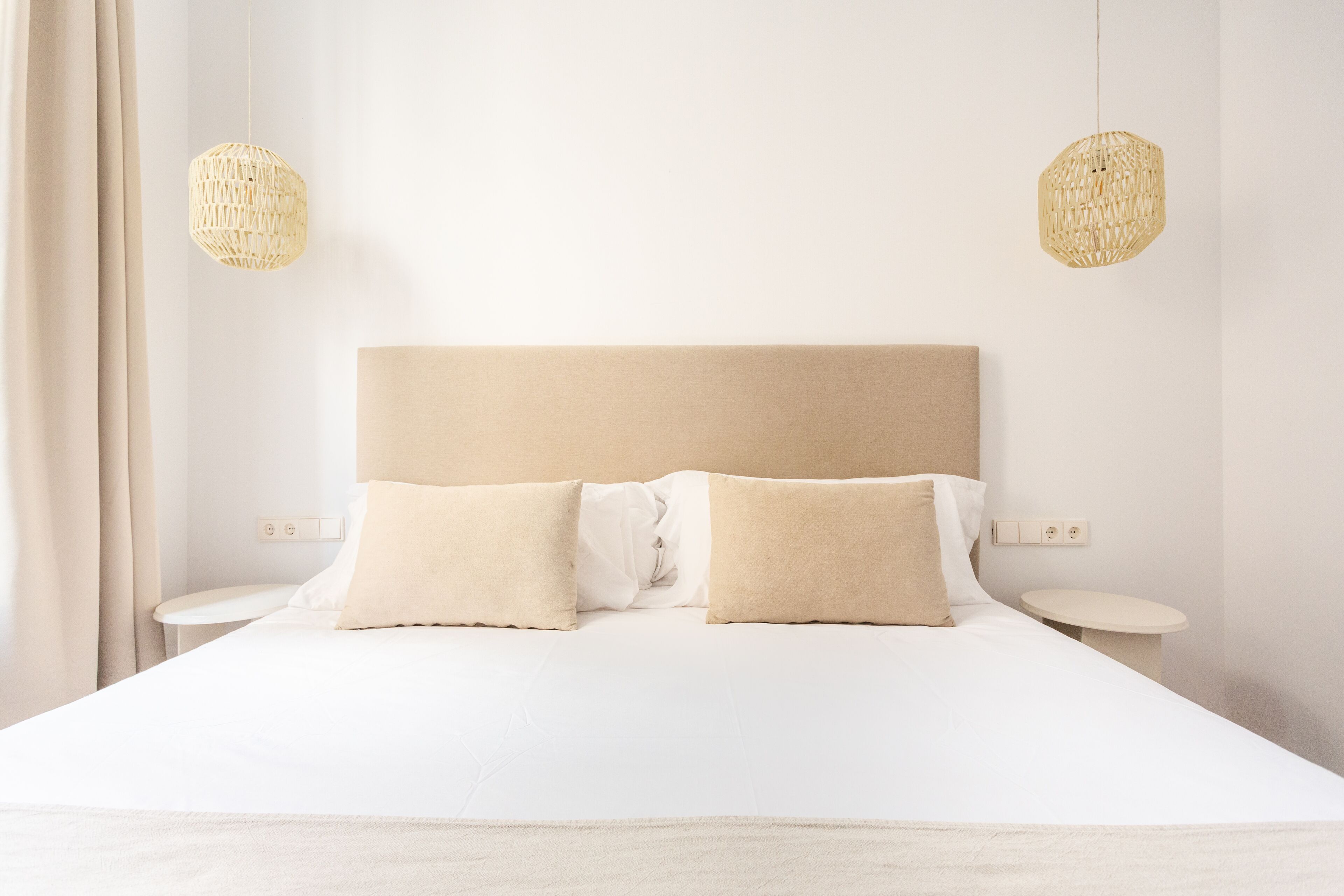 Una cama bien hecha con un cabecero color beige, dos mesitas de noche y dos lámparas colgantes tejidas.