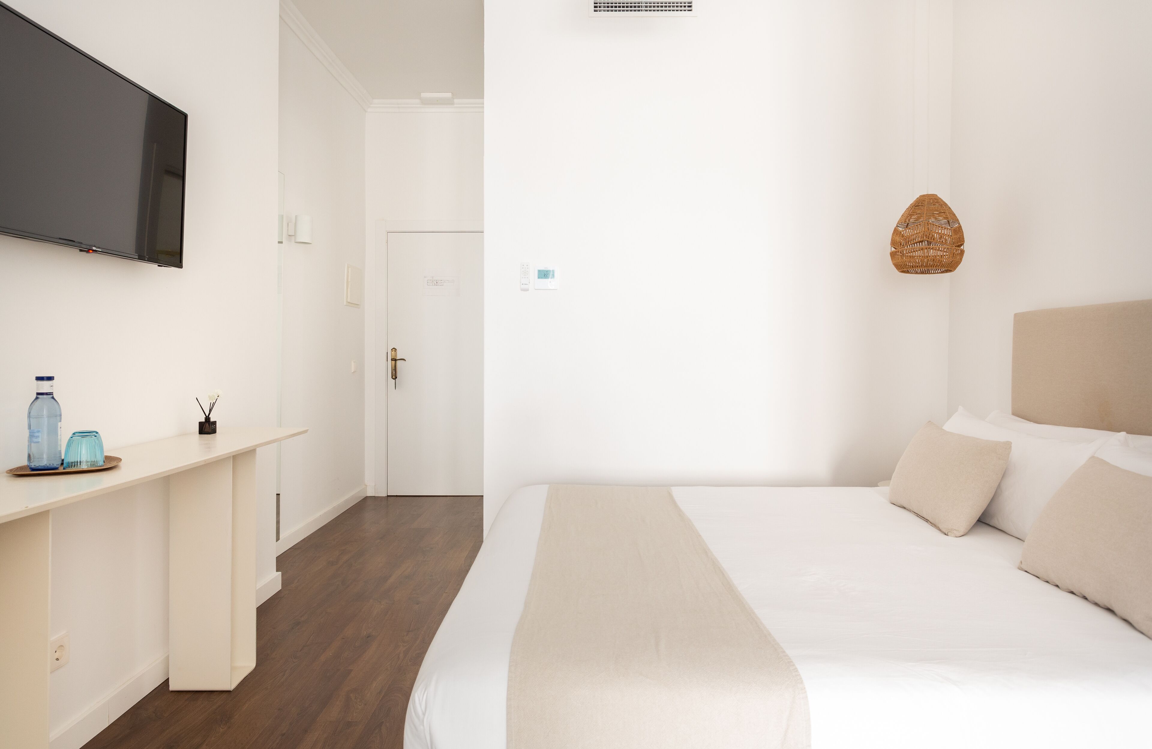 A hotel room with a bed, a TV mounted on the wall, a desk with a chair, and a door leading to another room.