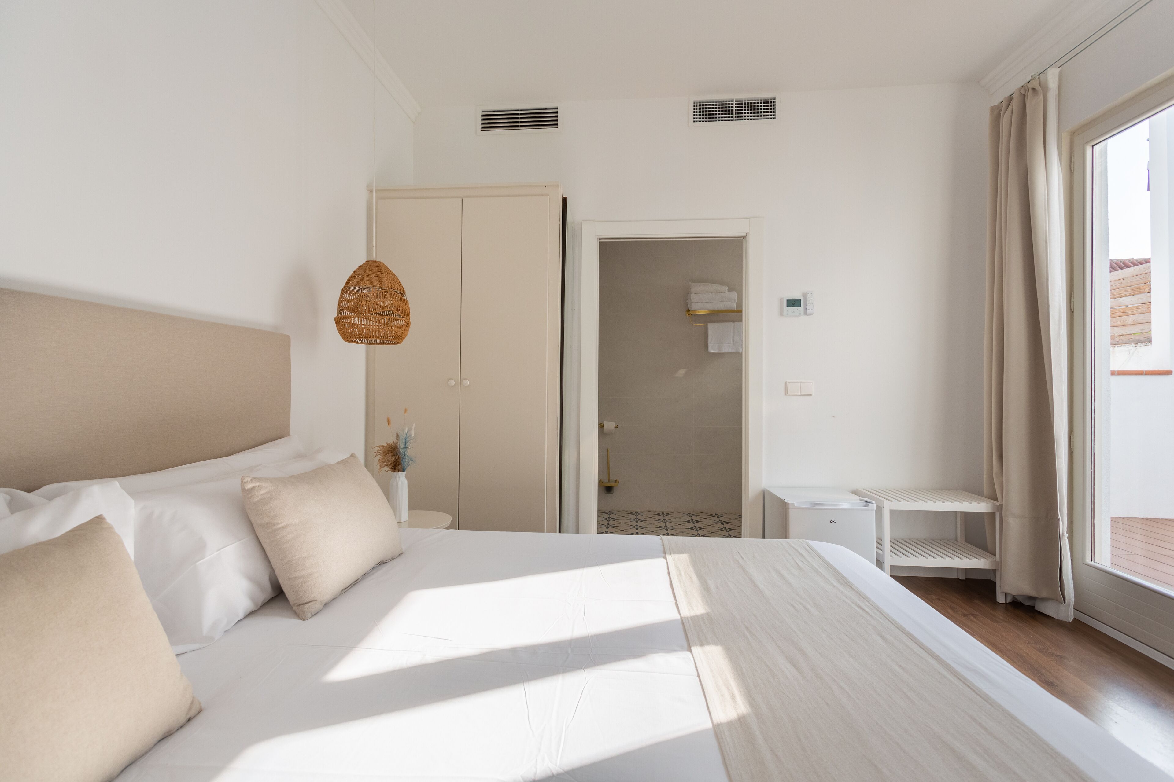 Un dormitorio con una cama grande, un armario, un baño visible a través de una puerta abierta y una mesita de noche pequeña.