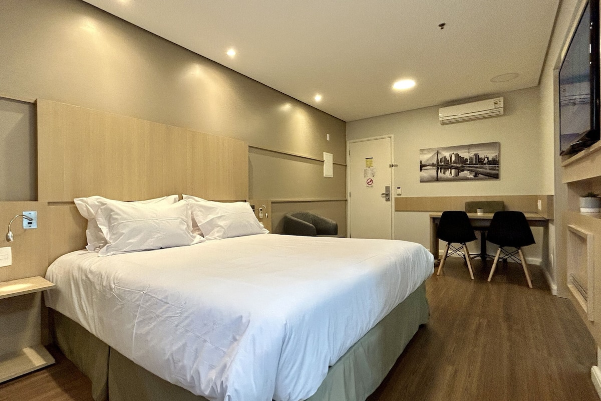 A hotel room with a large bed, a desk with two chairs, a mirror, and a wall-mounted air conditioner.