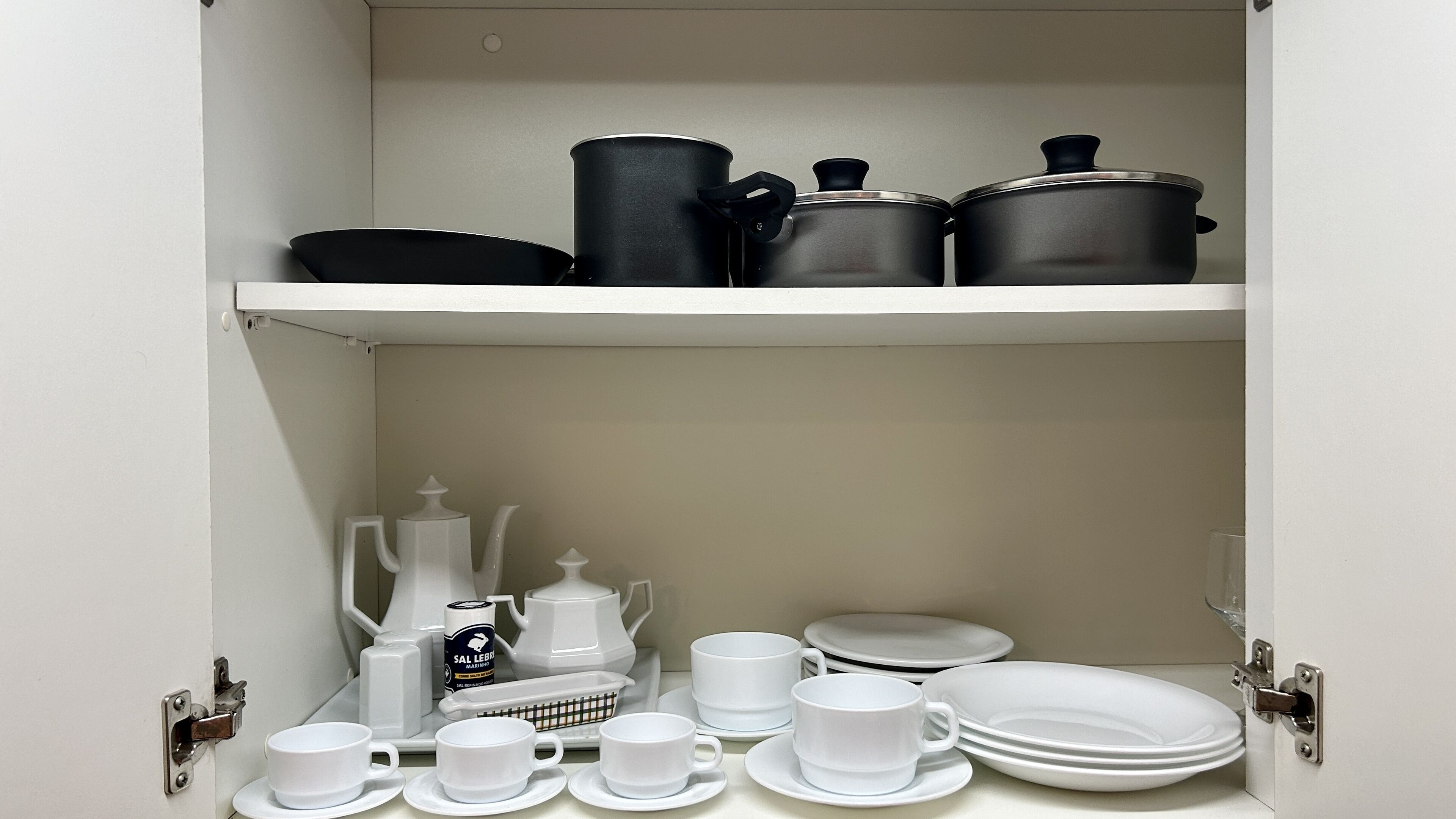 A kitchen cabinet with black and white cookware, a coffee maker, and white dishes.
