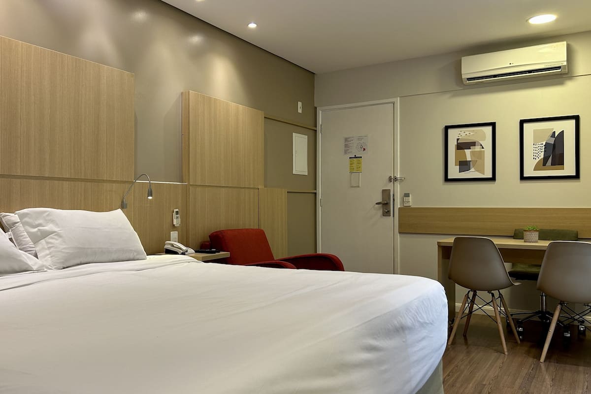 A hotel room with a large bed, a desk, a chair, and an air conditioning unit.