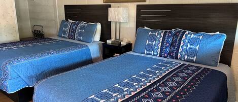 A hotel room with two beds, each with blue bedding and patterned pillows. There is a framed painting on the wall above the beds.