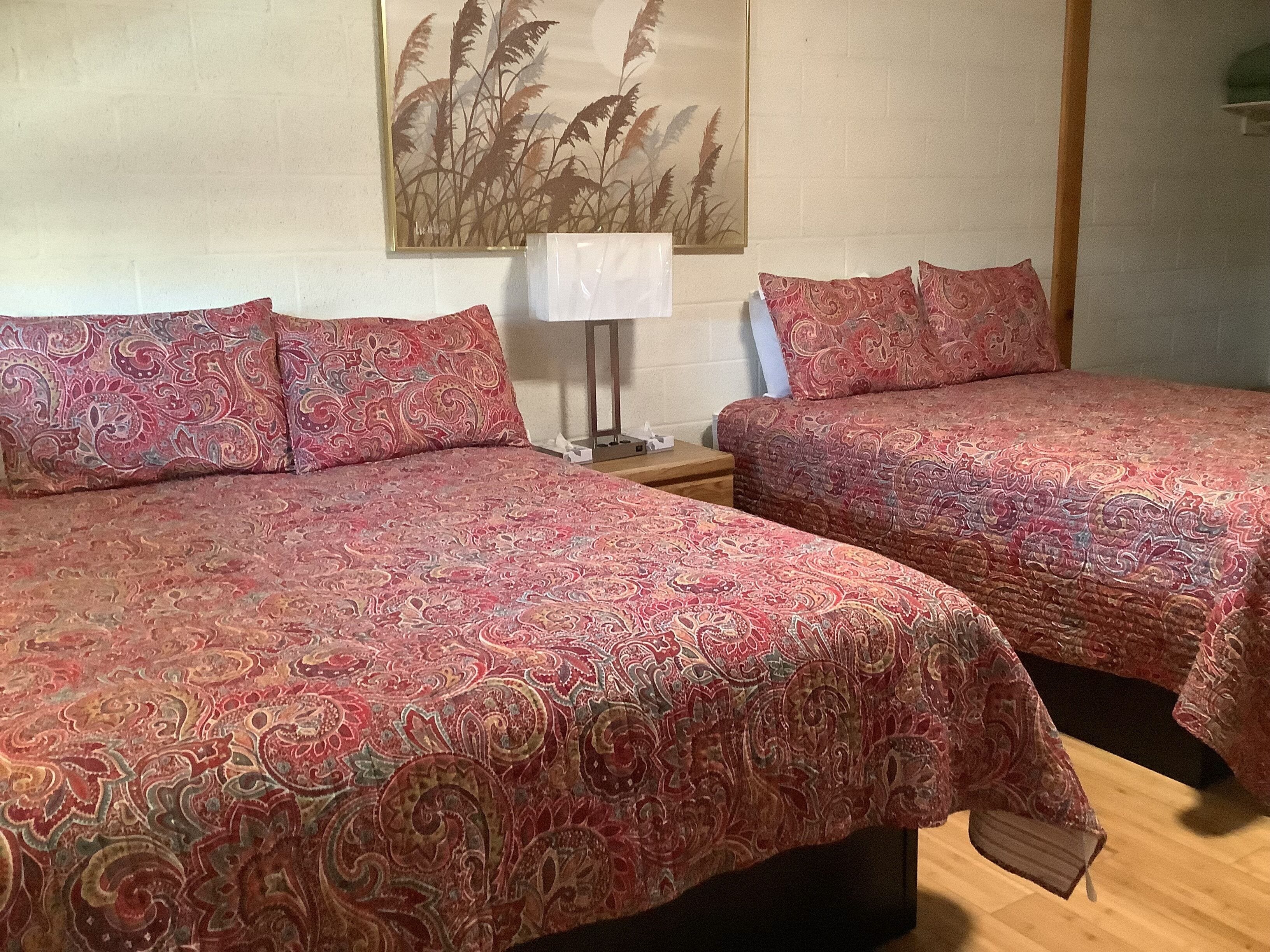 Quarto de hotel com duas camas, cada uma com colcha estampada, uma mesa de cabeceira com abajur e uma obra de arte pendurada na parede.