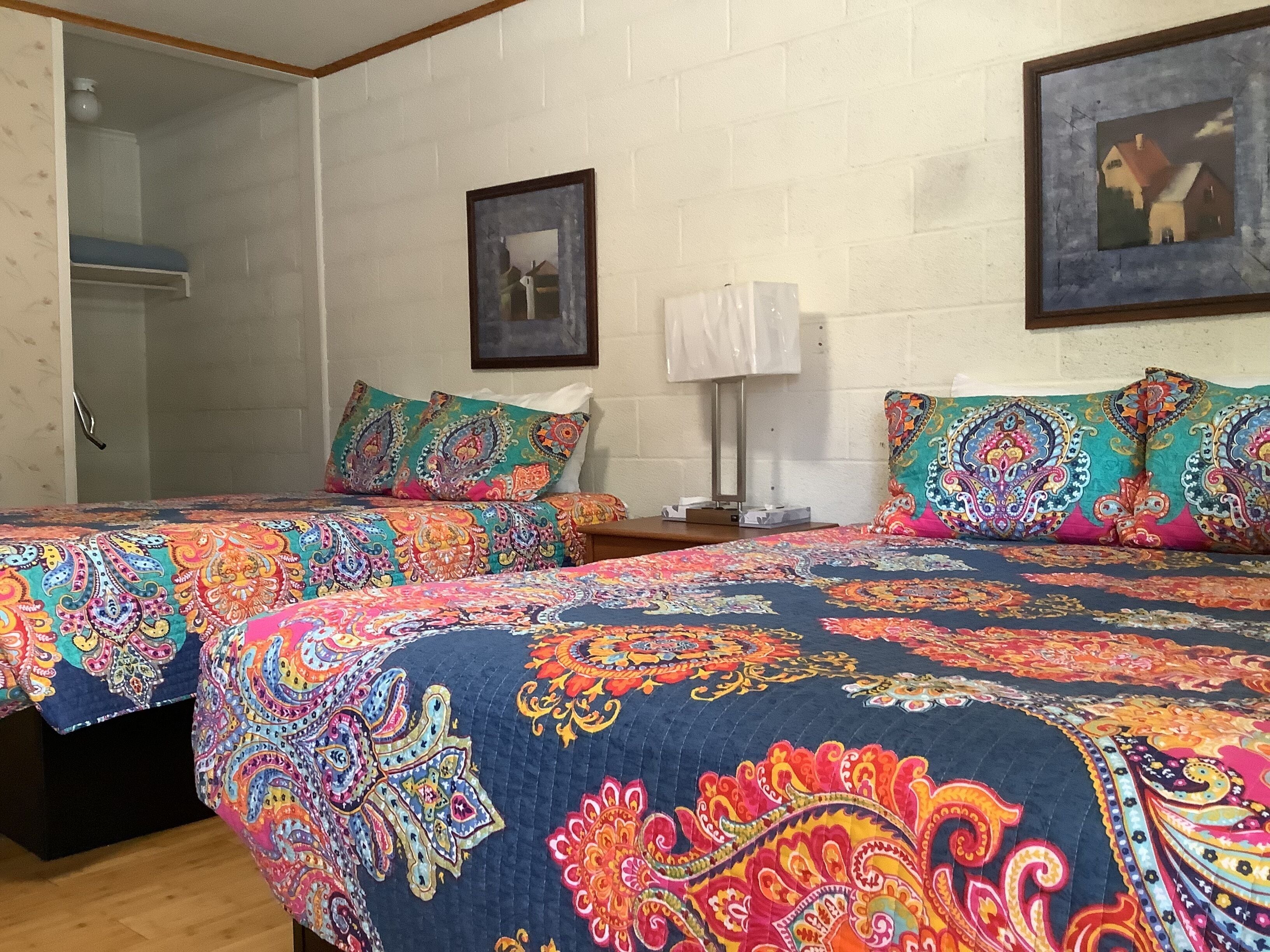 Una habitación con dos camas, cada una con ropa de cama de colores y diseños, una pared de ladrillo blanco, un cuadro enmarcado y una mesita de noche con lámpara.