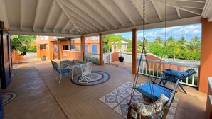 A covered patio with a table and chairs, a swing, and a view of a residential area with palm trees. - Walk to the beach (Christiansted)