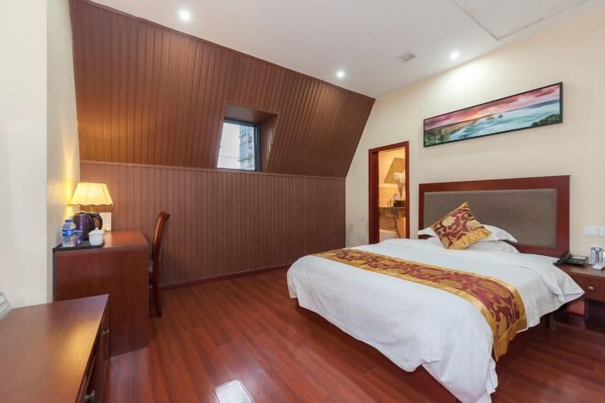 A hotel room with a wooden ceiling, a bed with a white and red bedspread, a painting on the wall, and a desk with a lamp.