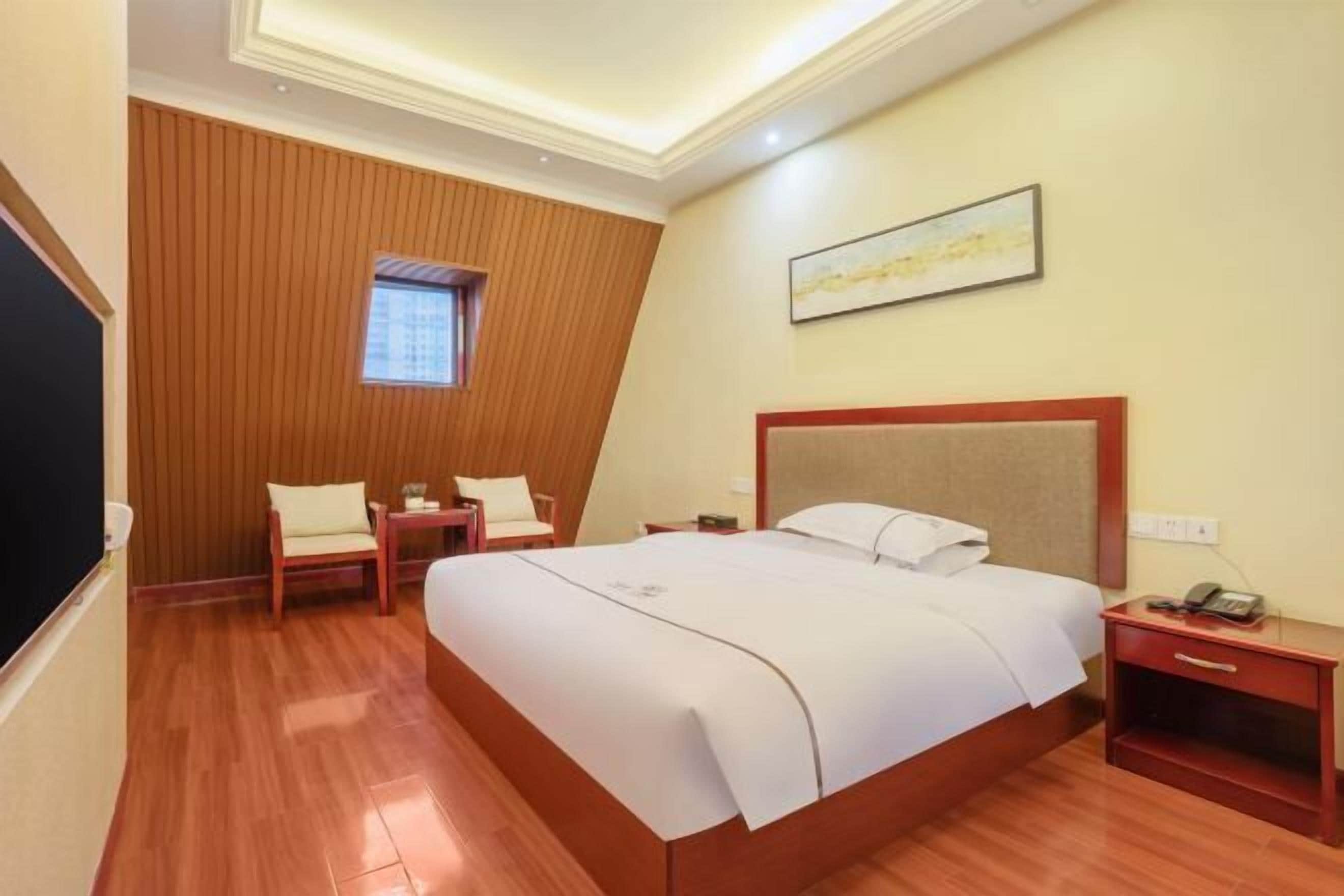 A hotel room with a large bed, a wooden accent wall, a small window, a nightstand, and a painting on the wall.