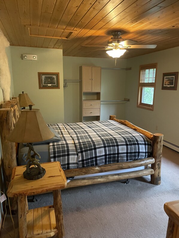 A wooden bed with a plaid blanket, a wooden nightstand with a lamp, a wooden chair, a wooden desk, a window with blinds, and a ceiling fan.