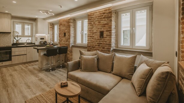 Un salon moderne avec un canapé beige, une petite table ronde et un coin cuisine doté d’un mur en briques et de grandes fenêtres.