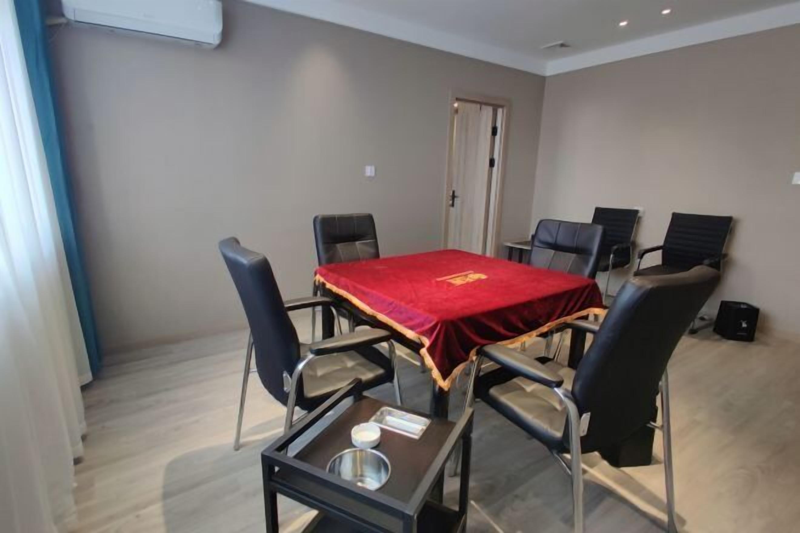 Un salón de conferencias con una mesa roja, seis sillas, una mesa pequeña con un bol y una servilleta, y una puerta al fondo.