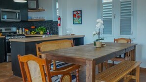 Une cuisine moderne dotée d’un îlot central, d’une table à manger en bois et de chaises. On y trouve des suspensions vertes, un extincteur et une fenêtre avec des volets.