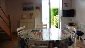 A dining area with a round table covered in a tablecloth, surrounded by white chairs. There is a staircase on the left, a door leading outside, and a television on the right wall. - House 5 minutes from the beach and shops (La Brée-les-Bains)