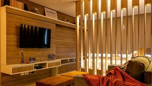 A modern living room with a flat-screen TV, wooden shelves, a bench, and a cozy seating area with a blanket. - 450m Shopping | Dining | Pool #CA25 (Florianópolis)