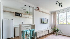 Een moderne keuken met een magnetron, koelkast en barkrukken. Er hangt een televisie aan de muur en er is een raam met gordijnen.