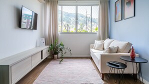Een moderne woonkamer met een bank, een klein bijzettafeltje, een aan de muur bevestigde televisie en een groot raam met gordijnen.
