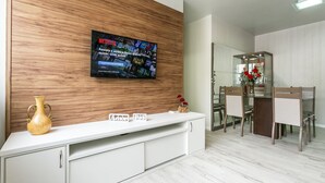 Een moderne woonkamer met een houten accentmuur, een flatscreen televisie, een witte console tafel, een vaas, en een eethoek met stoelen en een glazen kast.
