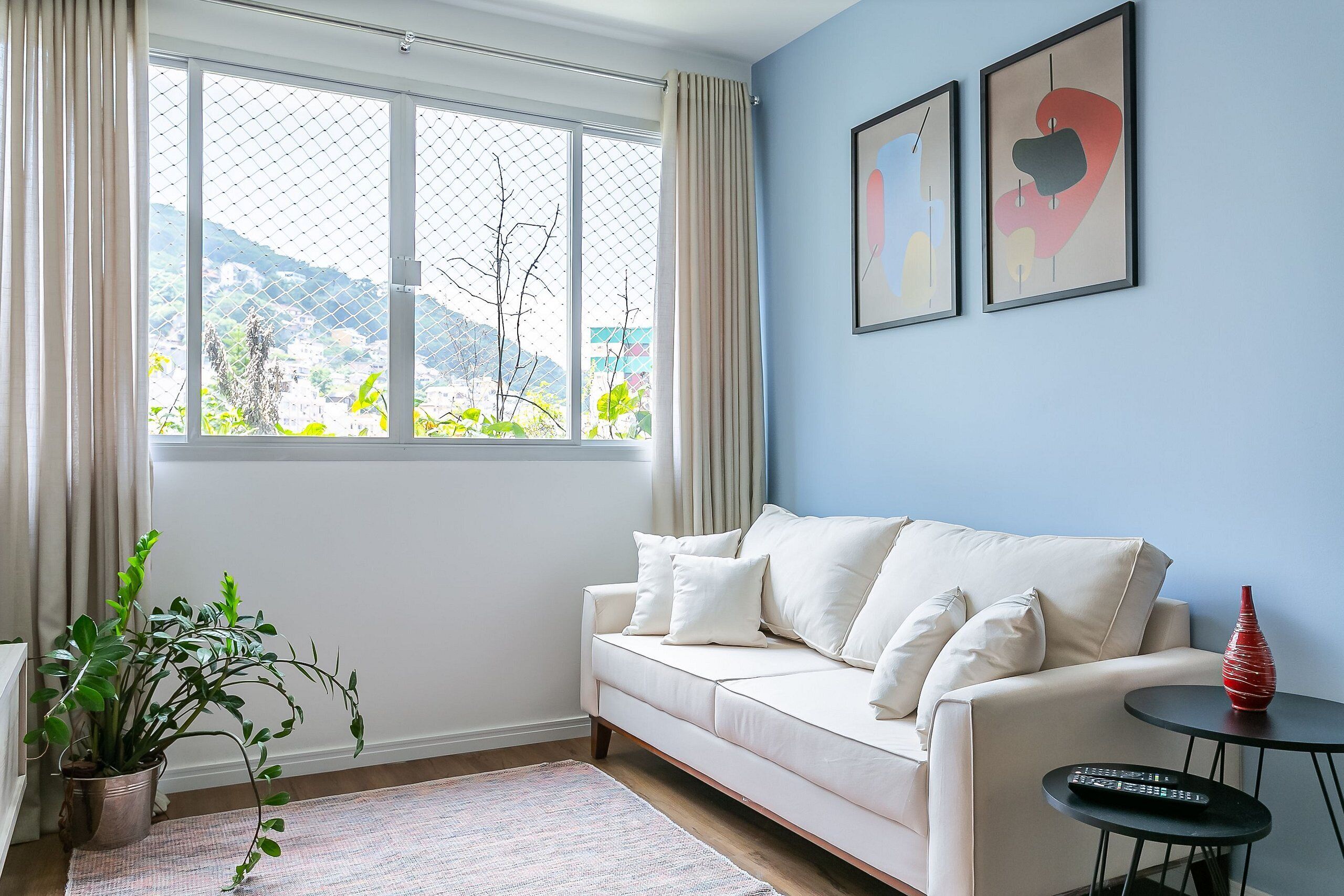 Ein Wohnzimmer mit einem weißen Sofa, einem kleinen Tisch mit einer Vase, einer Topfpflanze und einem Fenster mit Vorhängen.