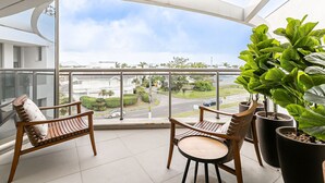 Ein Balkon mit zwei Holzstühlen und einem Tisch, großen Kübelpflanzen und Blick auf eine Straße und Gebäude.