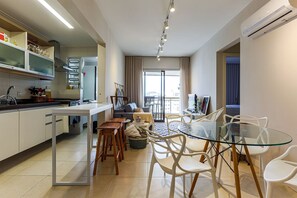 A modern kitchen with white cabinets, a dining area with a glass table and white chairs, and a living area with a sofa and a TV.