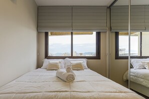 A neatly made bed with a rolled towel, two pillows, a window with blinds, and a mirror reflecting the bed. - WI-FI 250MB | Condominium with Pool #CA10 (Florianópolis)