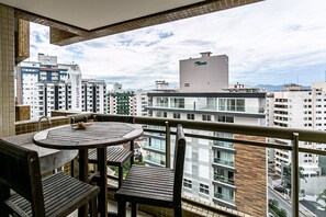 A balcony with a round wooden table and chairs, overlooking a cityscape with high-rise buildings and a clear sky.