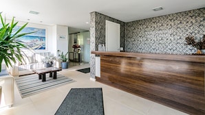 A hotel reception area with a wooden reception desk, patterned wall tiles, a large potted plant, and a sofa with striped cushions. - SEA VIEW | Balcony with Barbecue #IA09 (Florianópolis)