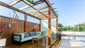 A covered outdoor area with a wooden deck, a light blue sofa, a small table, and a pool in the background. - SEA VIEW | Balcony with Barbecue #IA09 (Florianópolis)