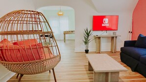 A modern living room with a wicker chair, a blue sofa, and a wooden coffee table. A red Nintendo Switch TV is mounted on the wall. - Fun Haven! 2 Units in 1 house for private gatherings and games (South Haven)