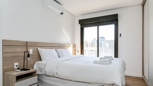Une chambre d’hôtel moderne avec une tête de lit en bois, un lit blanc doté d’un matelas matelassé, une table de chevet avec une lampe et une vue sur la ville par une grande fenêtre.