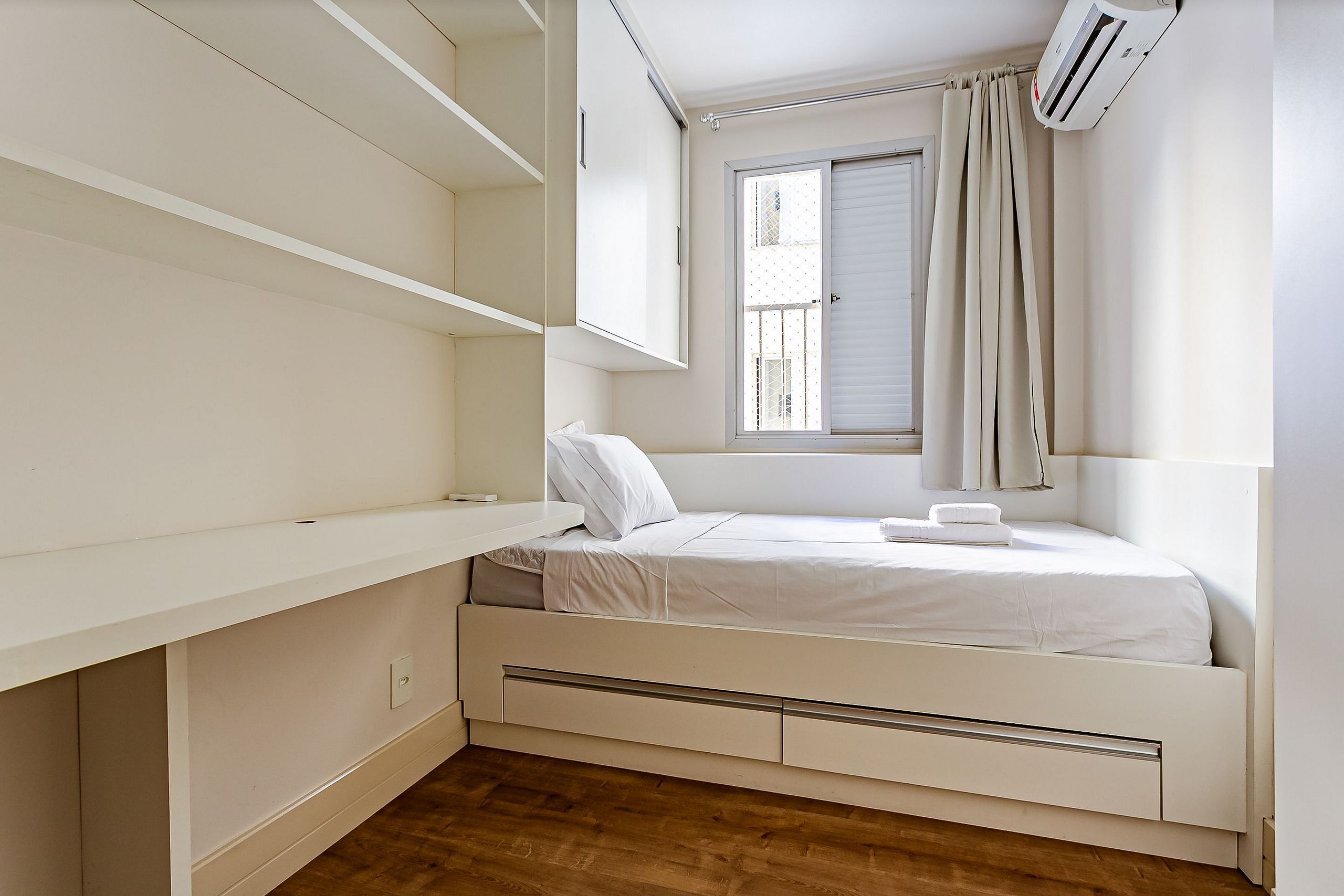 A minimalist bedroom with a bed, built-in storage, a window with curtains, and an air conditioning unit.