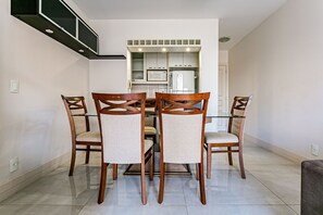 Uma área de refeições com mesa de vidro e cadeiras de madeira, uma cozinha com eletrodomésticos brancos e um chão ladrilhado.