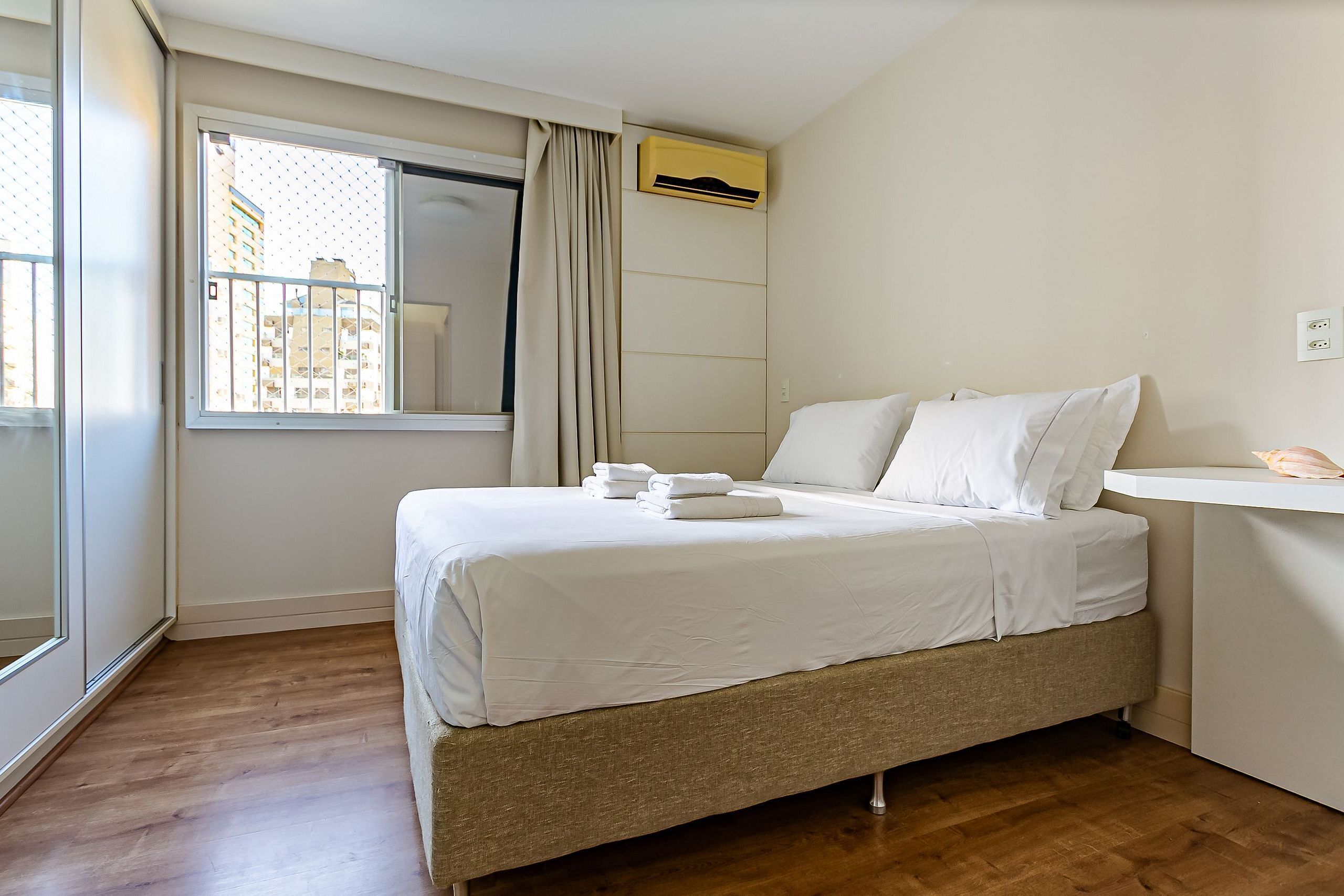 A neatly made bed with white linens, a window with a view of buildings, and a wall-mounted air conditioner.