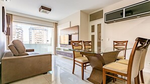 A modern living room with a glass dining table, wooden chairs, a sofa, and a TV mounted on the wall. - Dining | Partial Sea View | Walkable #CA28 (Florianópolis)