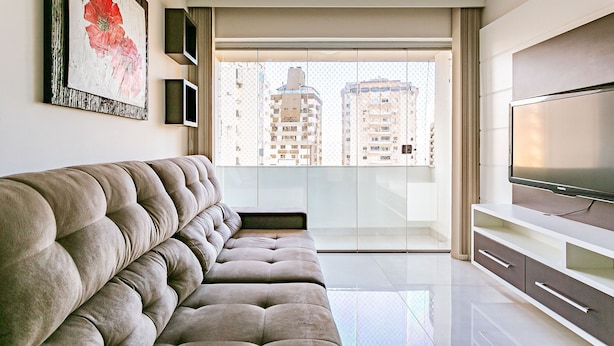 A modern living room with a tufted sofa, a flat-screen TV, and a large window offering a view of high-rise buildings. - Dining | Partial Sea View | Walkable #CA28 (Florianópolis)