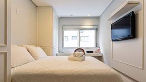 A neatly made bed with a quilted cover, a flat-screen TV mounted on the wall, and a window with blinds. - WI-FI 240MB | Downtown Florianópolis #CA19 (Florianópolis)