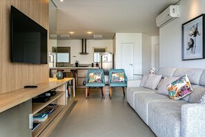 A modern living room with a grey sofa, teal chairs, a flat-screen TV, and a kitchen in the background.