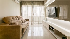 Uma sala de estar com sofá bege, televisão de tela plana e uma janela grande com cortinas finas.