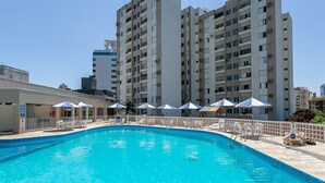 Piscina com espreguiçadeiras e guarda-sóis, cercada por uma grade, com edifícios residenciais altos ao fundo.