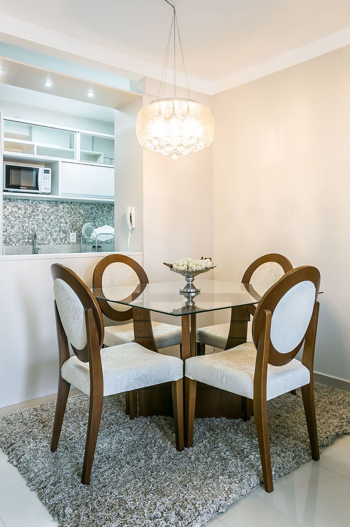 Un coin repas avec une table en verre entourée de quatre chaises, un lustre et un espace cuisine visible à travers une porte ouverte.