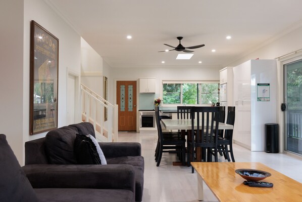 A modern living room with a sofa, a dining table with chairs, a kitchen area with a window, and a ceiling fan. - Anais Hat Head 5 Bedroom House 10 Guests (Hat Head)
