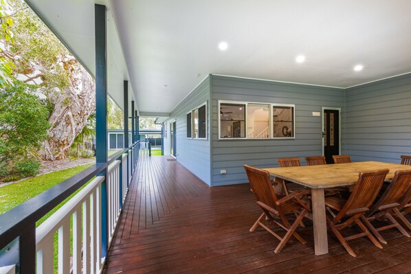A covered outdoor area with a wooden deck, a dining table with chairs, and a view of a garden and building in the background. - Anais Hat Head 5 Bedroom House 10 Guests (Hat Head)