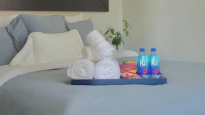 A neatly made bed with a blue bedspread, white pillows, and rolled towels. There are two bottles of Fiji water and a small plant on the side. - Bright Upper-Level 3BR Chicago • King • Walkable (Chicago)