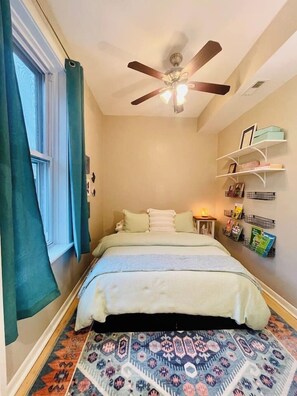 A bedroom with a bed, a ceiling fan, a window with curtains, and a wall with shelves holding books and decorative items.