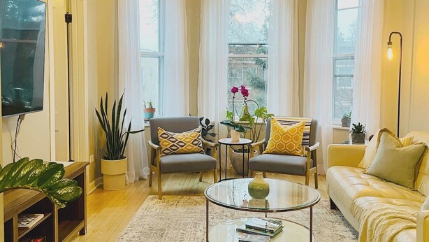 A cozy living room with a sofa, two armchairs, a coffee table, and a TV. There are potted plants and a window with curtains. - Bright Upper-Level 3BR Chicago • King • Walkable (Chicago)