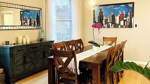 A dining area with a wooden table and chairs, a dark-colored cabinet, a window with white curtains, and a cityscape painting on the wall. - Bright Upper-Level 3BR Chicago • King • Walkable (Chicago)