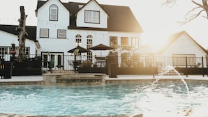 Ein Poolbereich mit einer Fontäne, ein weißes Gebäude mit zahlreichen Fenstern und Gartenmöbeln.