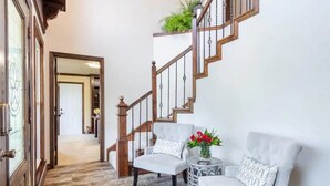 Un escalier avec des rampes en bois et un mur blanc. On y trouve deux fauteuils blancs avec des coussins à motifs et une petite table ronde sur laquelle est posé un vase de fleurs.