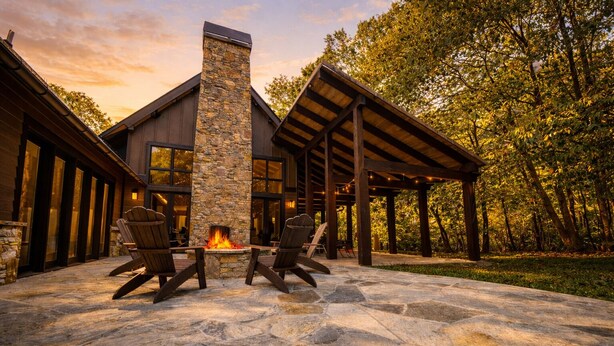 A stone patio with a fire pit and two Adirondack chairs, surrounded by a wooden pergola and a stone chimney.