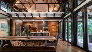 A spacious dining area with a long wooden table, modern kitchen in the background, and large windows offering a view of the outdoors.